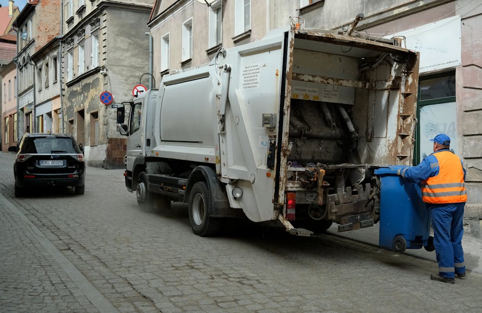 A black wheelie bin with the label 'ST. JOHN'S' positioned on a paved sidewalk beside a street at night. The bin is filled with various discarded items including cardboard boxes, plastic packaging, an